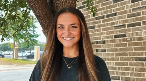 Katie Harnen smiling while standing in front of a tree with a brick wall in the background